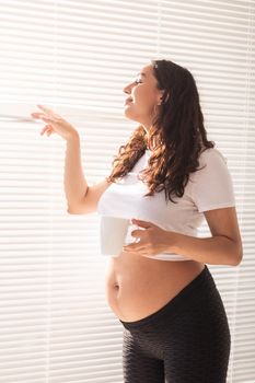Curious young beautiful pregnant woman drinking tea and looking through the blinds at the window. Joy and good news while waiting for baby