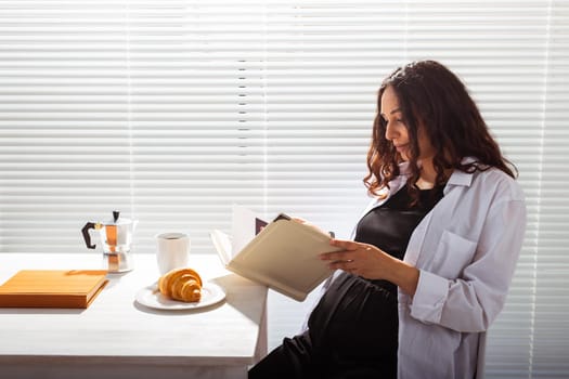 Side view of happy young beautiful woman reading book while having morning breakfast with coffee and croissants on background of blinds. Good morning and pleasant lunch break