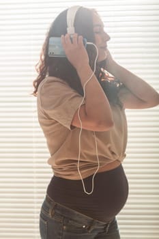 Curly-haired brunette pacified pregnant woman listens to pleasant classical music using smartphone and headphones. Concept of a soothing mood before meeting baby