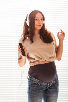 Young peaceful pregnant woman listens to pleasant classical music using smartphone and headphones. Concept of positive attitude before childbirth