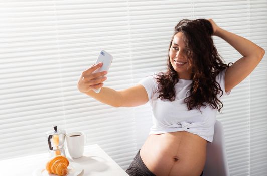 Pregnant woman takes selfie on her smartphone while breakfast. Maternity leave and technologies