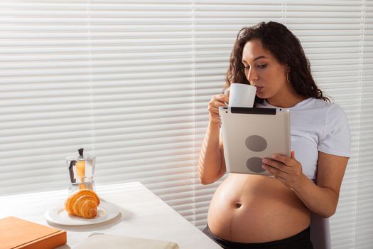 Hispanic pregnant woman using digital tablet while breakfast. Technology, pregnancy and maternity leave.