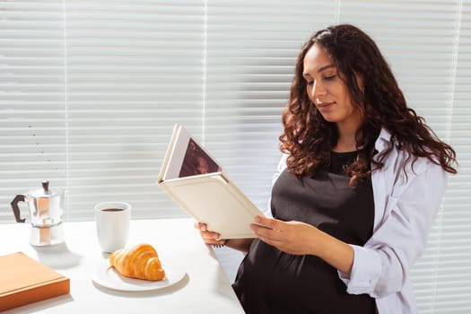 Side view of happy young beautiful woman reading book while having morning breakfast with coffee and croissants on background of blinds. Good morning and pleasant lunch break