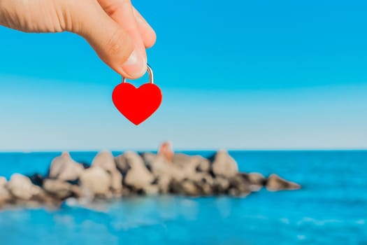 The man's hand holds a small red heart in the form of a castle against the backdrop of the sea coast and the stones of the breakwater on the horizon line. Symbol or sign of love.