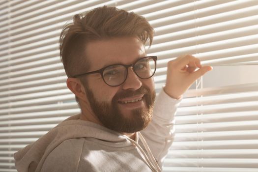 Rear view of young man with beard peeks through hole in the window blinds and looks out into the street. Surveillance and curiosity