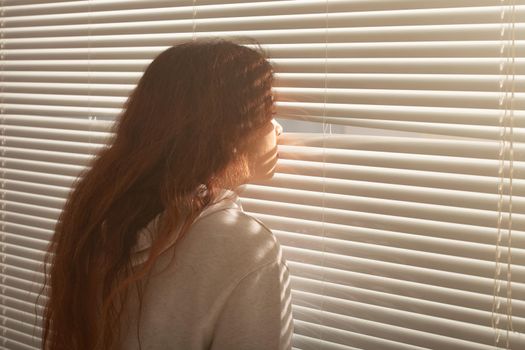 Rear view of beautiful young woman with long hair peeks through hole in the window blinds and looks out the window. Surveillance and curiosity