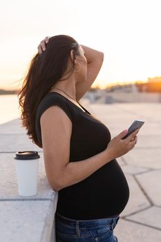 Pregnant woman with long hair holds smartphone outdoors. Side view. Warm day. New life, new horizons and opportunities