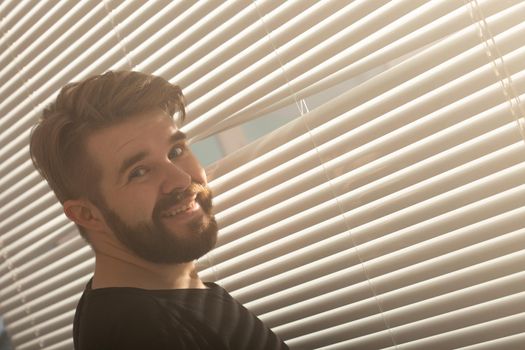 Rear view of young man with beard peeks through hole in the window blinds and looks out into the street. Surveillance and curiosity
