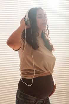 Curly-haired brunette pacified pregnant woman listens to pleasant classical music using smartphone and headphones. Concept of a soothing mood before meeting baby