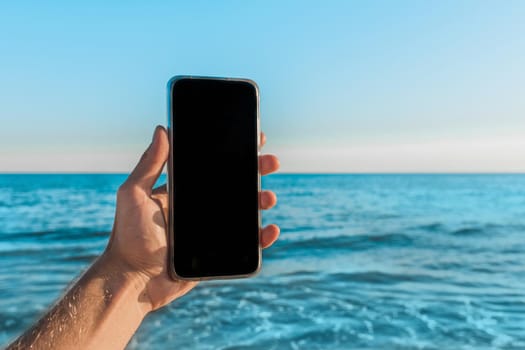 The man's hand holds a mobile phone with a black screen space for text and design against the blue sea, sky and skyline.