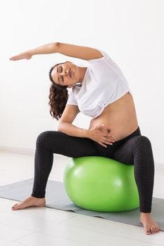 A young pregnant woman doing relaxation exercise using a fitness ball while sitting on a mat and holding her tummy