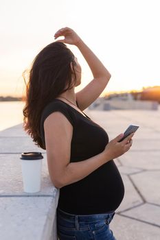 Pregnant woman with long hair holds smartphone outdoors. Side view. Warm day. New life, new horizons and opportunities