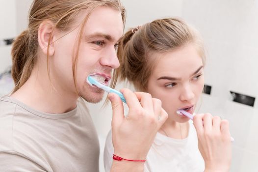 Cute young couple in the morning in a white bath brush their teeth together with toothbrushes, looking at the reflection in the mirror, laughing and smiling. Close-up