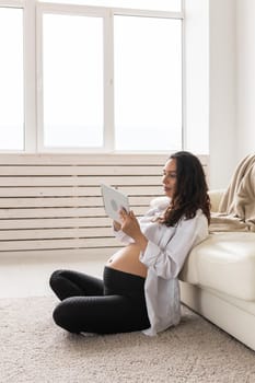 Pregnant woman holding tablet sitting on a carpet near a couch in the living room at home.