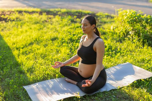 Cute young caucasian pregnant woman is meditating while sitting on a rug on the lawn on a sunny summer evening. Concept of pacification