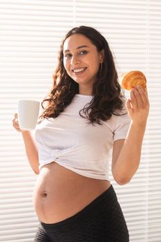 Beautiful pregnant woman holding croissant and cup of coffee in her hands during morning breakfast. Good health and positive attitude while expecting baby