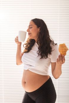 Pregnant woman eating croissant and drinks coffee. Pregnancy and maternity leave.