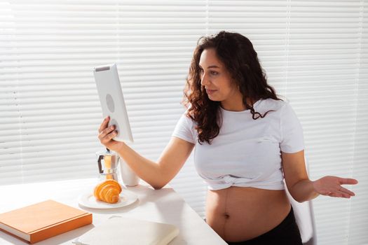 Happy pregnant young beautiful woman talking to mom using video call during morning breakfast. Communication and positive attitude during pregnancy