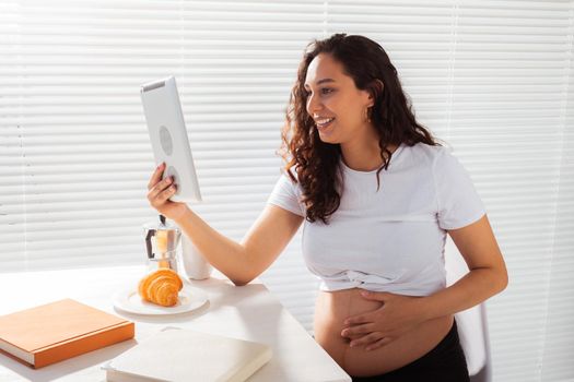 Happy pregnant young beautiful woman talking to mom using video call during morning breakfast. Communication and positive attitude during pregnancy