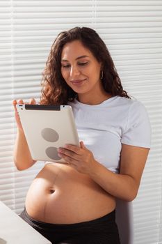 Hispanic pregnant woman using digital tablet while breakfast. Technology, pregnancy and maternity leave.