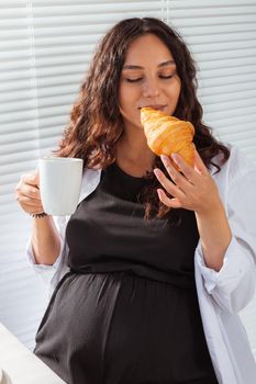 Happy pregnant young beautiful woman eating croissant and looking througt blinds during morning breakfast. Pleasant morning and positive attitude during pregnancy