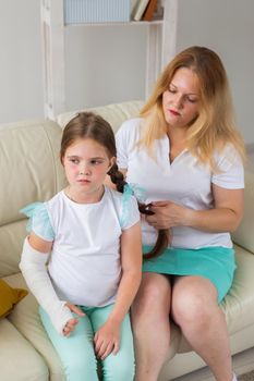 Mother braids a braid your child girl with a cast on a broken wrist or arm smiling. Childhood illnesses, a positive outlook and recovery