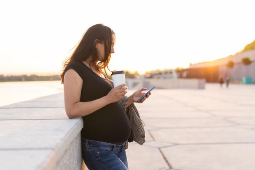 Young pregnant woman drinking takeaway coffee or tea on embankment. Maternity, leisure and health