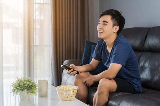 cheerful young man holding remote control and watching TV while sitting on sofa in the living room 