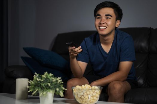 cheerful young man holding remote control and watching TV while sitting on sofa at night