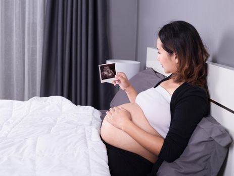 pregnant woman with ultrasound image of baby sitting on bed in the bedroom at home