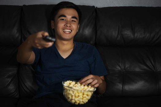 cheerful young man holding remote control and watching TV while sitting on sofa at night