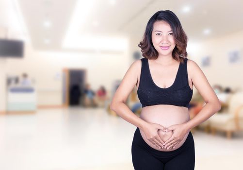 Pregnant Woman holding her hands in a heart shape on her belly in hospital background