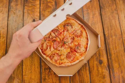 The guy's hand opens a cardboard box of pizza against the background of a wooden table. Delicious fast food.