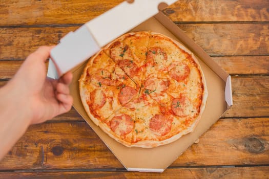 The guy's hand opens a cardboard box of pizza against the background of a wooden table. Delicious fast food.