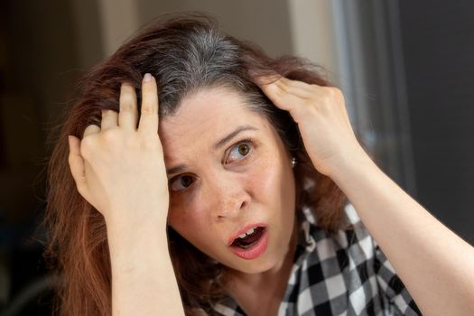 Horrified young woman looking in the bathroom mirror staring open mouthed at the first grey hair on her scalp, a first sign of ageing, or noticing that she is suffering from dandruff