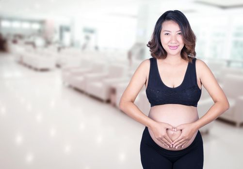 Pregnant Woman holding her hands in a heart shape on her belly in hospital background