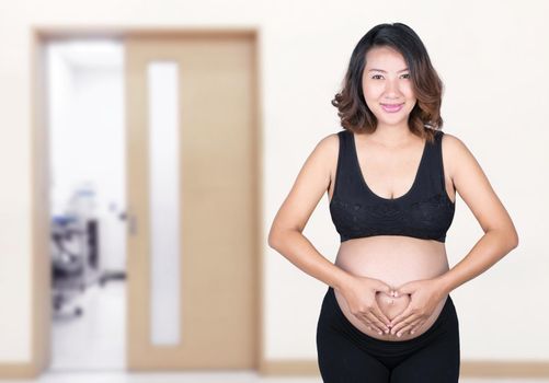 Pregnant Woman holding her hands in a heart shape on her belly in hospital background