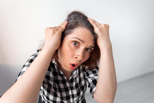 Horrified young woman looking in the bathroom mirror staring open mouthed at the first grey hair on her scalp, a first sign of ageing, or noticing that she is suffering from dandruff