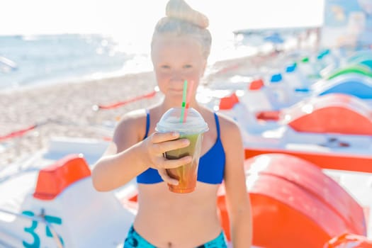 A young girl with blonde hair of European appearance, a teenager holds a colored cold non-alcoholic cocktail in her hand against the background of the sea beach.