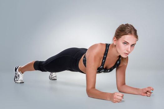 Sporty fit beautiful young woman in sportswear bra and black pants working out, doing plank pose for abdominal muscles, studio full length, gray background. Sports lifestyle