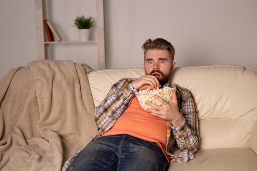 Bearded man watching film or sport games TV eating popcorn in house at night. Cinema, championship and fan.
