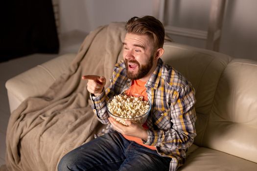 Portrait young man sitting on couch and watching TV holding remote and eats popcorn
