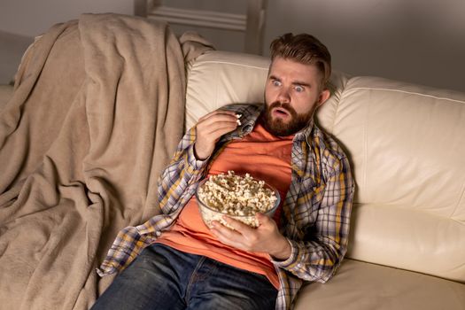 Bearded man watching film or sport games TV eating popcorn in house at night. Cinema, championship and fan.
