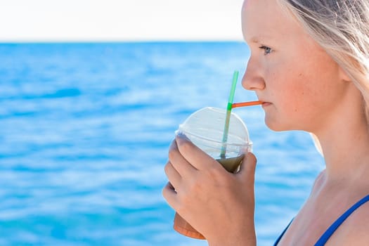 A young girl with blonde hair of European appearance, a teenager holds and drink a colored cold non-alcoholic cocktail in her hand against the background of the sea beach.