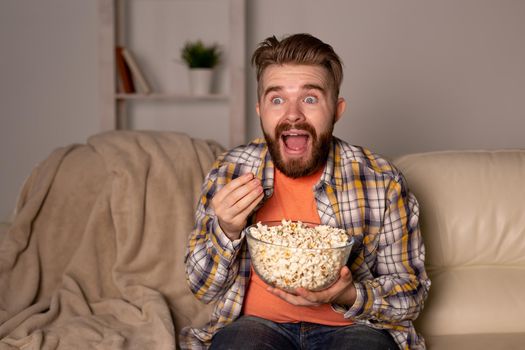 Bearded man watching film or sport games TV eating popcorn in house at night. Cinema, championship and fan.