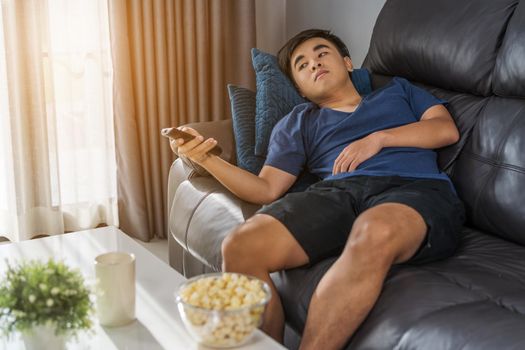 young man holding remote control and watching TV while sitting on sofa in the living room 