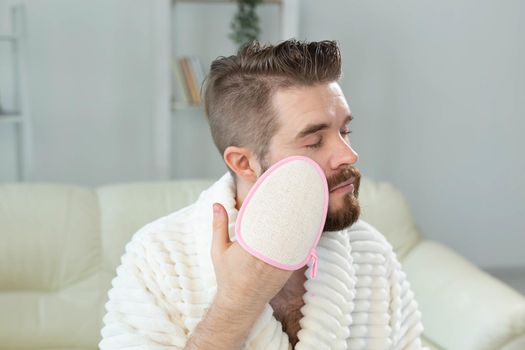 Bearded man look in mirror and clean scrub face with sponge, happy millennial male metrosexual do beauty procedures at home, take care of healthy skin, skincare concept.