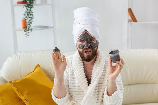 Bearded man having fun with a cosmetic mask on his face made from black clay. Men skin care and spa at home.