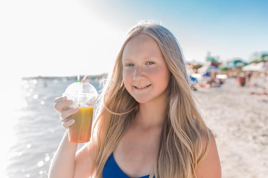 Young, cheerful attractive girl blonde of European appearance teenager in a blue swimsuit with a colored non-alcoholic cocktail in his hand against the background of the sea beach and the seashore.