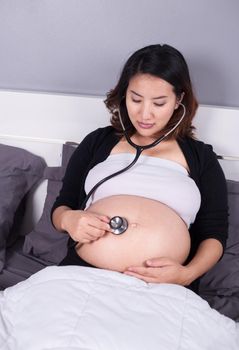 Pregnant woman listening to her belly with a stethoscope on the bed
